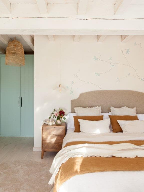 Dormitorio principal luminoso con cabecero tapizado en beige, cojines en tonos tostados y blancos, y un delicado mural pintado a mano con ramas y pájaros en azul. El armario verde menta y la lámpara de mimbre añaden calidez natural al espacio.
