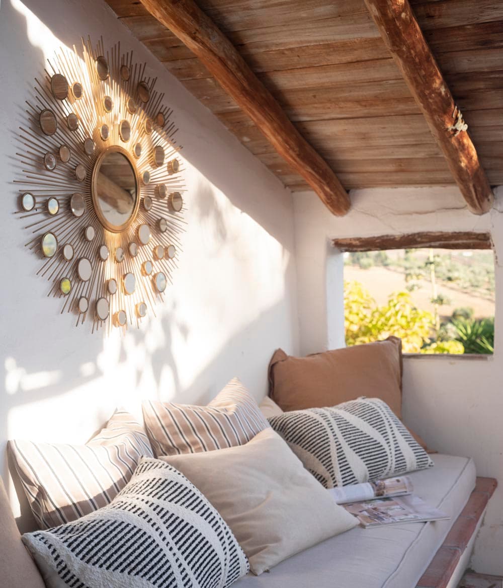 Acogedor rincón de descanso con banco corrido de obra tapizado con cojines de rayas y estampado geométrico, bajo un espejo sol dorado. El techo de vigas de madera natural y una pequeña ventana con vistas al jardín completan el ambiente mediterráneo.