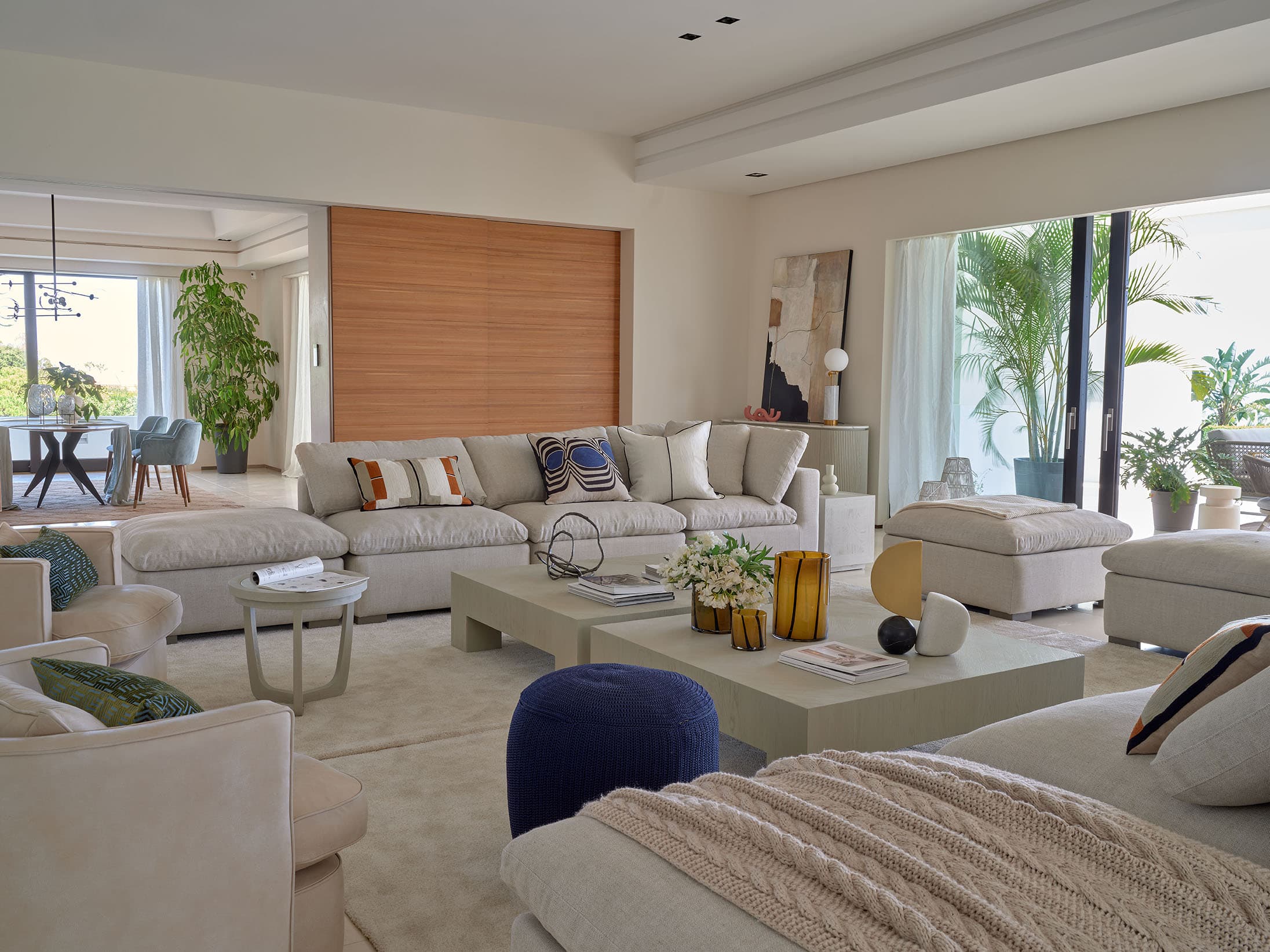 Salón de tonos cálidos y neutros con sofá modular profundo en lino beige, cojines con estampados abstractos, puf azul marino, mesas de café lacadas y acceso visual al comedor.