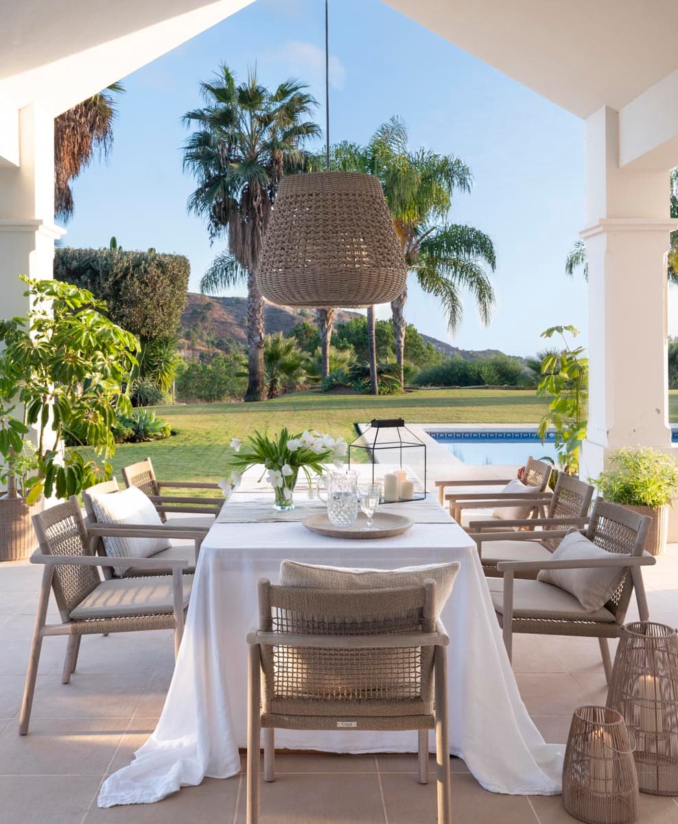 Elegante comedor exterior bajo porche con mesa rectangular cubierta de mantel blanco y sillas de cuerda trenzada en gris. Una gran lámpara de cuerda colgante preside el espacio abierto al jardín tropical con palmeras, piscina azul y montañas al fondo. Farol y candelabro añaden atmósfera al conjunto.
