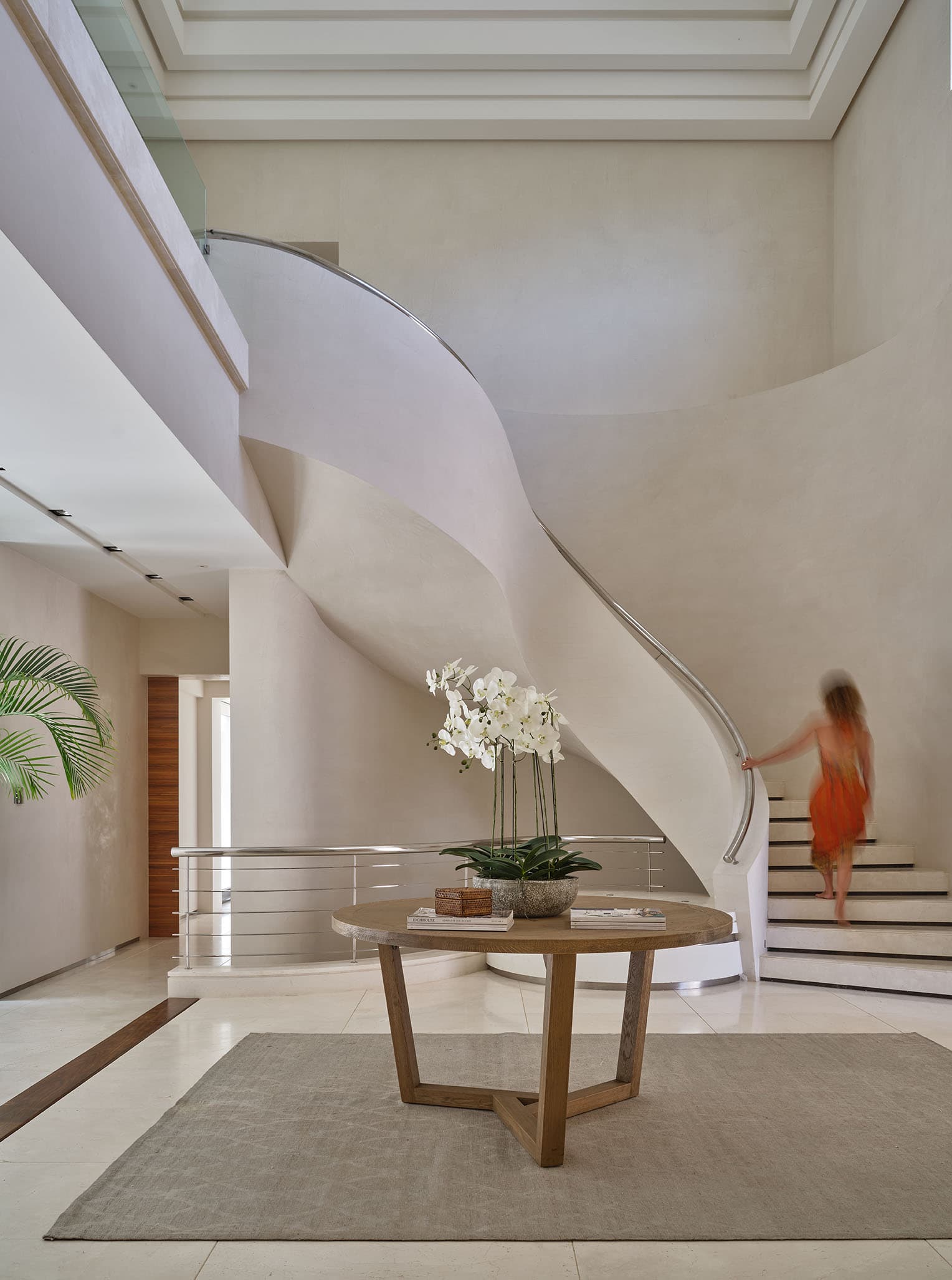 Impresionante hall de entrada de una villa en La Zagaleta con escalera helicoidal de gran altura en piedra caliza y pasamanos de acero, con una mesa redonda central con orquídeas.