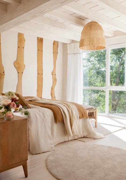 Dormitorio con vigas de madera natural en la pared a modo de cabecero artístico, ropa de cama en lino beige y ochre, y un gran ventanal que inunda el espacio de luz natural y vistas al jardín. Lámpara colgante de rafia y alfombra redonda completan el estilo rural elegante.