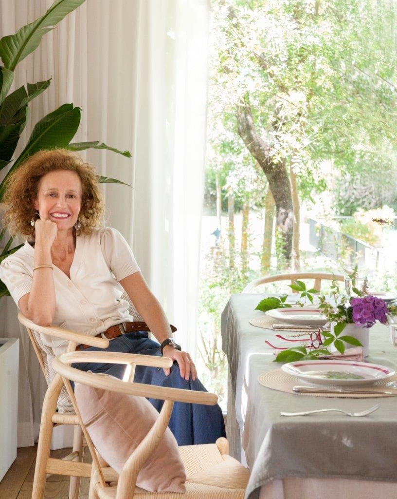 Retrato de la diseñadora de interiores Dafne Vijande sentada en una silla Wishbone de madera natural junto a la mesa del comedor decorada con flores. Vestida con una blusa blanca y pantalón vaquero, posa con naturalidad frente al ventanal con vistas al jardín verde. Un ambiente de elegancia sencilla y femenina.