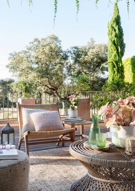 Terraza exterior con suelo de madera de teca y vistas al jardín rodeado de cipreses y encinas. Un sillón bajo de madera y ratán con cojín de lino y una mesa de mimbre circular sirven como zona de relajación. En primer plano, una mesa de mimbre con jarrones de vidrio verde y plantas caladium rosas.