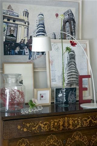 Detalle del aparador isabelino con acercamiento a las obras de arte urbano que representan edificios emblemáticos de Madrid, lámpara de arco articulada de metal blanco, tarro de cristal con rama coral roja y flor solitaria. La mezcla de estilos y épocas define el carácter coleccionista y culto del hogar.