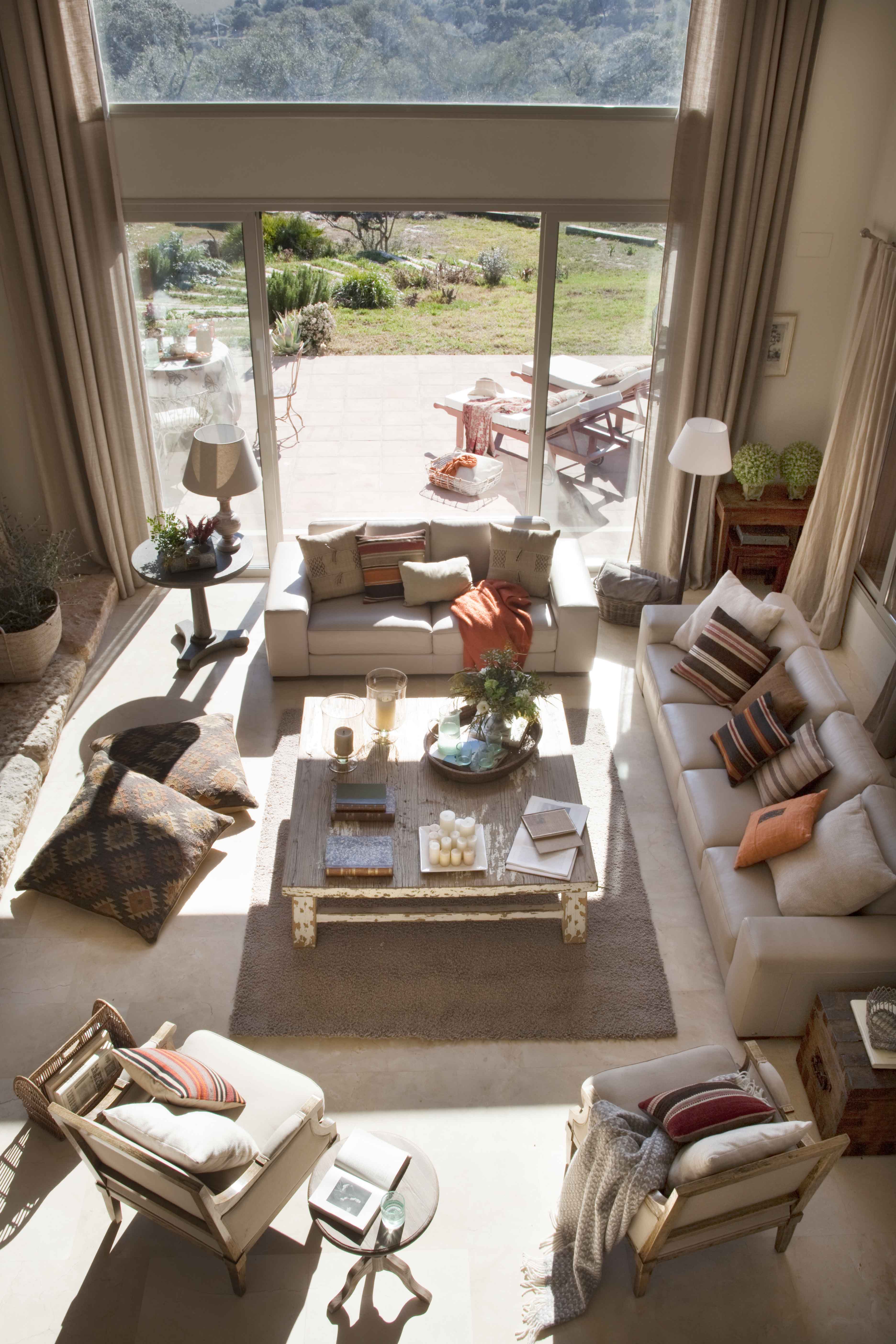 Vista cenital del amplio salón que se abre hacia una terraza exterior y jardín con vistas al campo. El espacio reúne múltiples zonas de estar con sofás en beige y crudo, cojines de kilim, una mesa central de madera envejecida y sillones de estilo francés. Los colores naranja y terracota de los almohadones aportan calidez al conjunto.