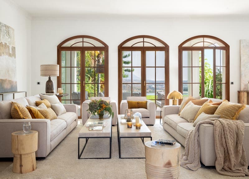 Salón amplio y sereno con ventanales en arco que enmarcan vistas a la montaña, sofás en lino beige con cojines en ocre y dorado, y una mesita de café de mármol con base de metal.