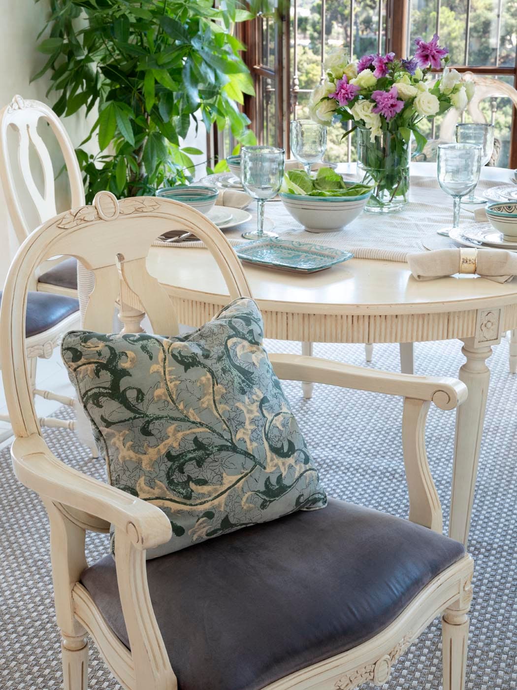 Primer plano de una silla de madera blanca estilo gustaviano con cojín de jacquard en azul verdoso, junto a una mesa redonda puesta con vajilla blanca, copas de agua y flores primaverales. La atmósfera informal y elegante es característica del estilo mediterráneo de la villa.