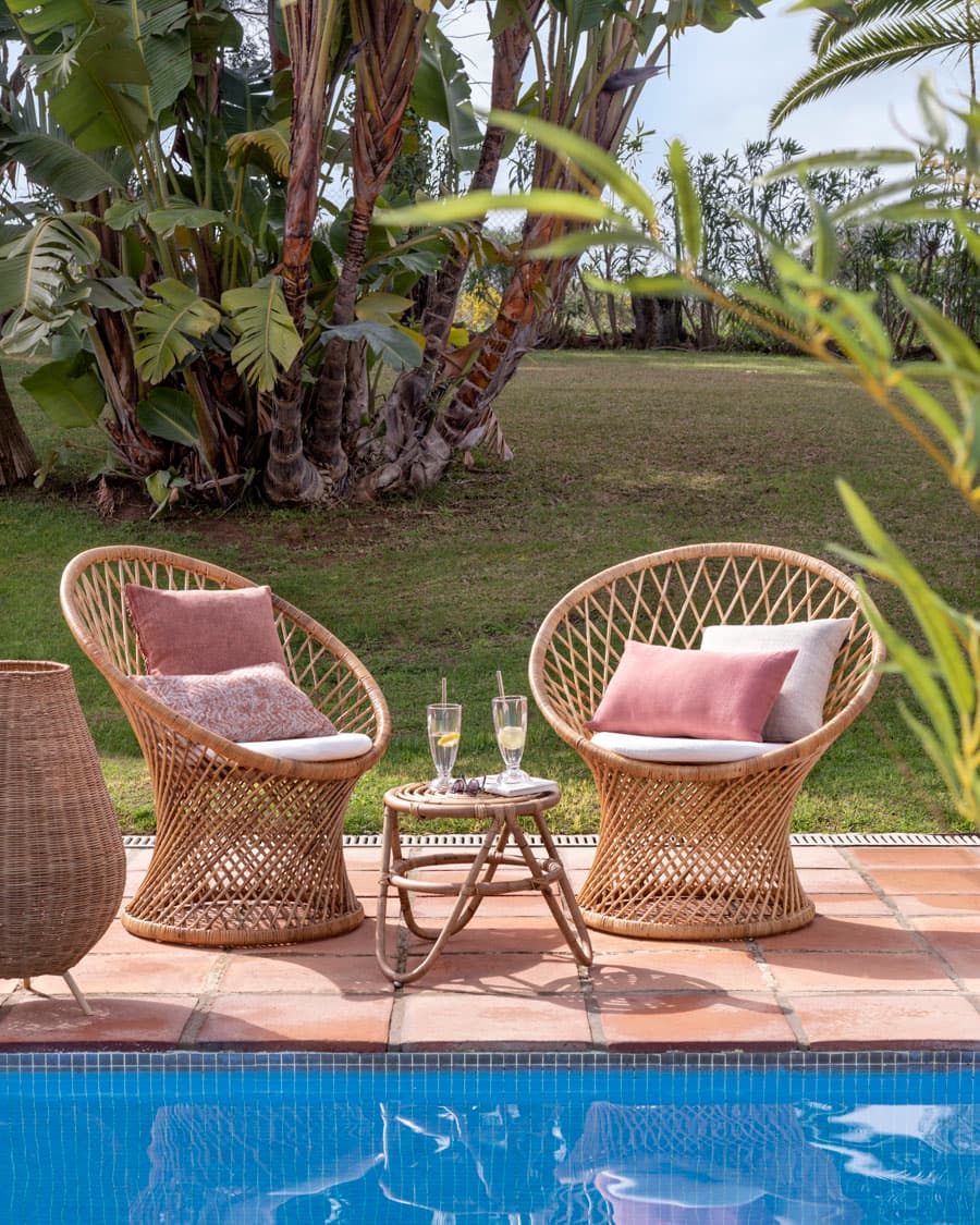 Zona de relax junto a la piscina con dos butacas de ratán con forma circular, cojines en rosa palo y malva suave, y una mesa auxiliar pequeña de ratán entre ellas. Sobre la mesa, dos copas de cava con manzanilla o cava, creando un ambiente de descanso y lujo. El entorno de jardín tropical con palmeras y monstera enmarcan la escena junto al borde de la piscina con suelo de barro cocido terracota.
