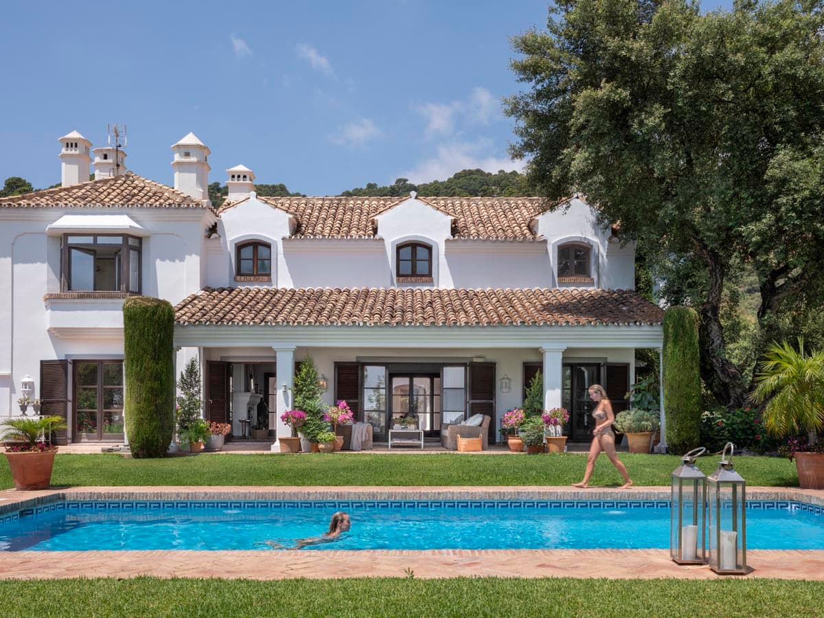 La misma fachada posterior de la villa capturada con bañistas en la piscina, transmitiendo el ambiente vibrante y familiar de la propiedad en verano. La arquitectura blanca y los jardines exuberantes refuerzan la imagen de villa de lujo andaluza en pleno disfrute.