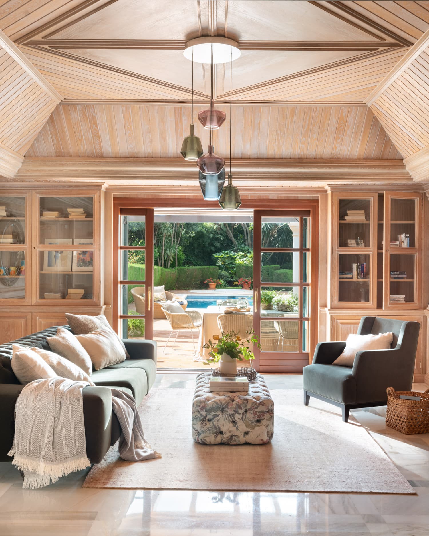 Sala de estar singular con techo abovedado de listones de madera natural en espiga, estanterías empotradas acristaladas y tres lámparas colgantes geométricas en tonos ahumados. Un sofá capitoné en verde oscuro y un sillón gris pizarra flanquean un puf de diseño floral. La puerta francesa abierta revela la terraza y la piscina del jardín.