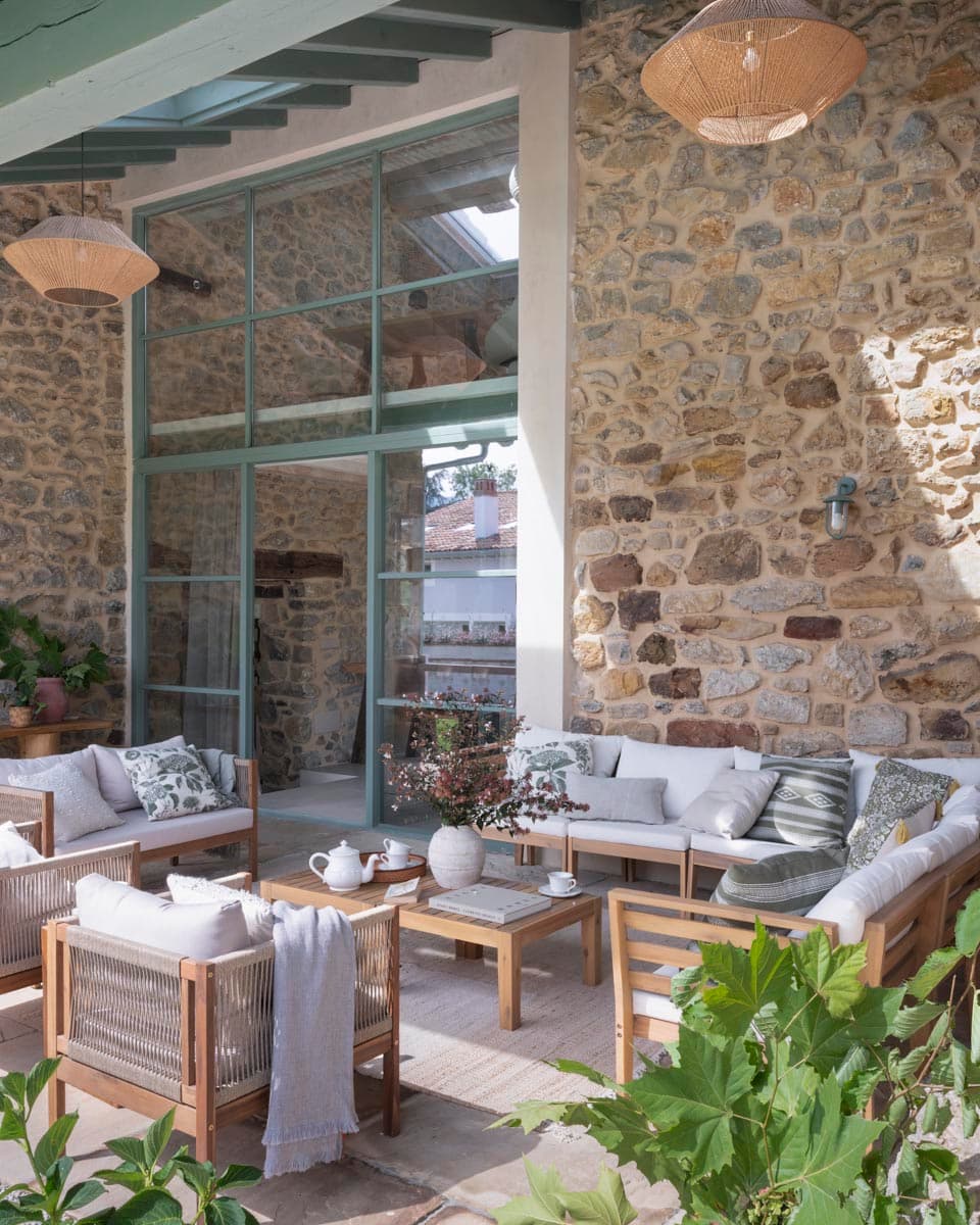 Porche cubierto con muro de piedra vista, sofás y butacas de madera con tapizado gris, lámparas de ratán colgantes y grandes ventanales acristalados verdes que dan acceso al interior luminoso.