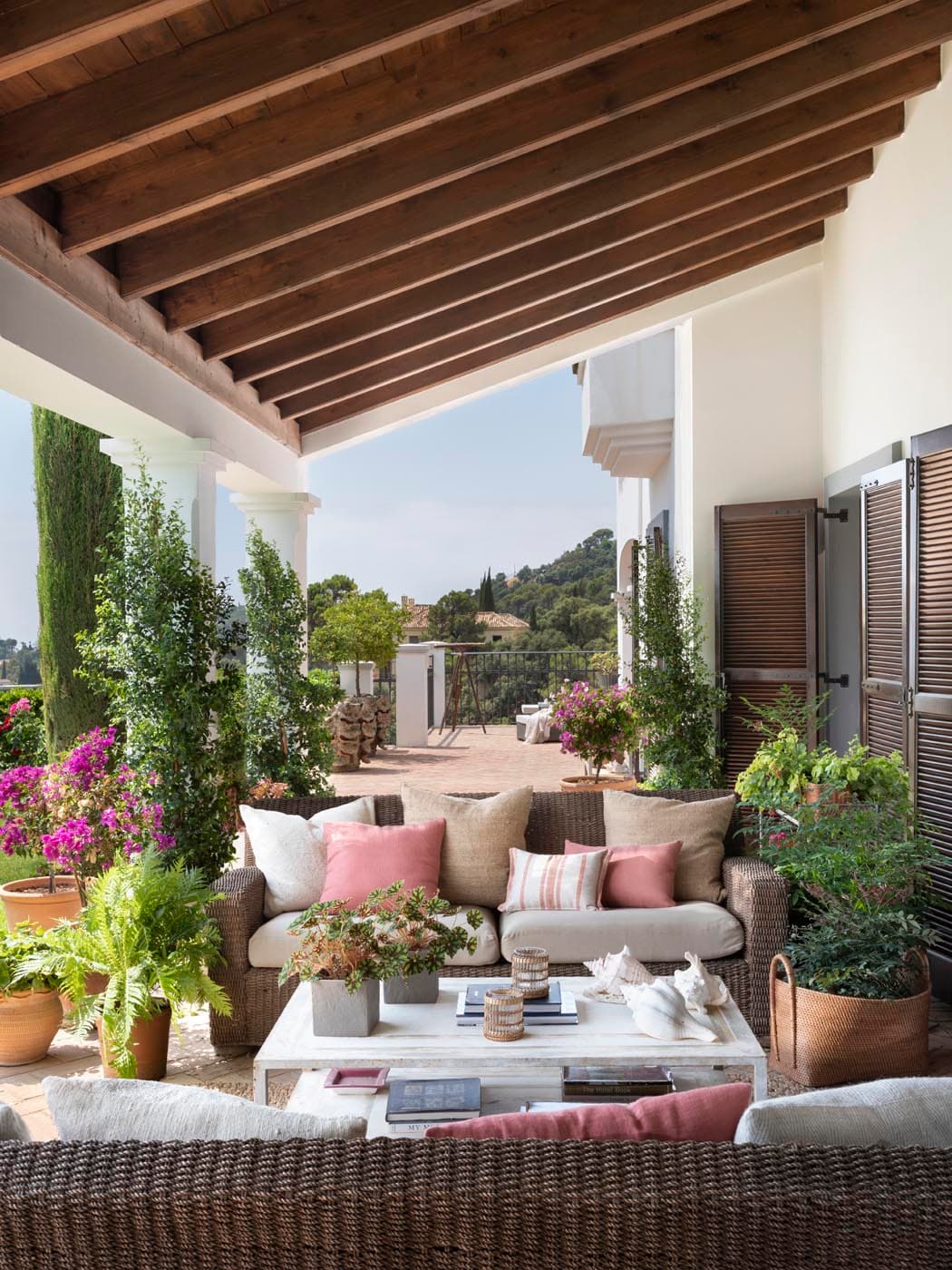 Amplia terraza cubierta con columnas blancas, vigas de madera y sofás de rattan con cojines en tonos rosados y arena, rodeada de exuberante vegetación mediterránea, buganvilias y cipreses. La vista hacia el jardín y la arquitectura encalada al fondo crean un ambiente de serenidad y lujo al aire libre.