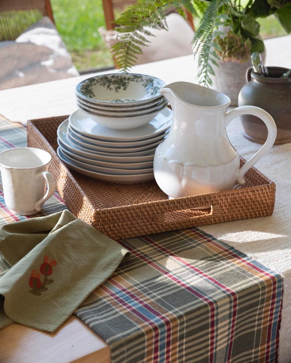 Detalle de bandeja rectangular de mimbre con platos y cuencos de porcelana blanca con borde verde, jarra blanca de cerámica y servilleta de lino verde con bordado floral, sobre camino de mesa a cuadros.