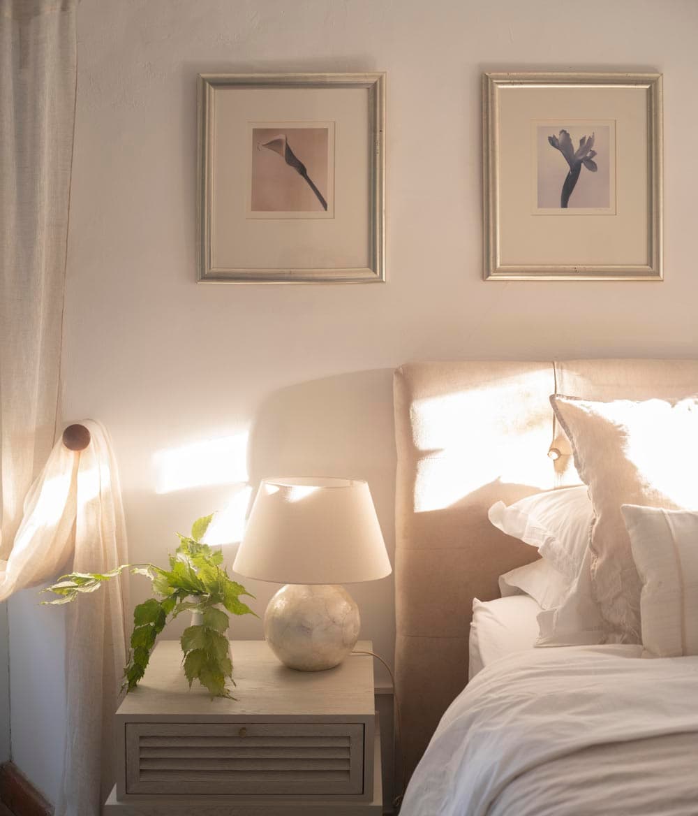 Detalle de cabecero tapizado en beige con mesita de noche, lámpara esférica de cerámica y dos cuadros botánicos enmarcados en plata sobre la pared blanca. La luz natural entra a través de una cortina de lino creando un ambiente cálido y sereno.
