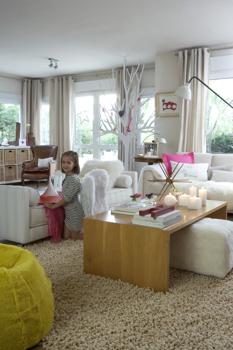 Vista del salón principal con decoración navideña, mostrando a una niña sonriente de pie junto al sofá blanco con cojín de piel. El árbol decorativo blanco con ornamentos festivos centra la estancia. La mesa de centro de madera de roble con velas, flores y libros ocupa el primer plano. Los sofás blancos de gran formato, el cojín fucsia vibrante, el puf amarillo y la lámpara de suelo configuran un ambiente acogedor y lleno de color. La luz natural entra generosamente por los grandes ventanales con cortinas de lino.