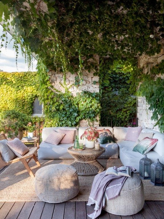 Porche exterior exuberante totalmente cubierto por hiedra y enredaderas que cuelgan del techo y trepa por las paredes de piedra. Sofá exterior blanco con cojines en rosa y verde, puf de fibra natural y silla de madera. Faroles marroquíes, cestas y manta lila crean un ambiente boho-mediterráneo.