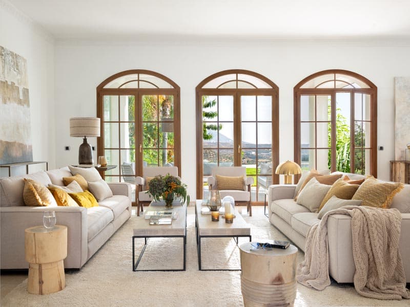 Salón principal con tres grandes ventanas en arco de carpintería de madera noble, sofás crema con cojines en mostaza y ocre, mesa de centro de travertino con estructura metálica y reposapiés de tronco. Los cuadros abstractos grandes a ambos lados y la lámpara de seta dorada equilibran el ambiente luminoso y contemporáneo.