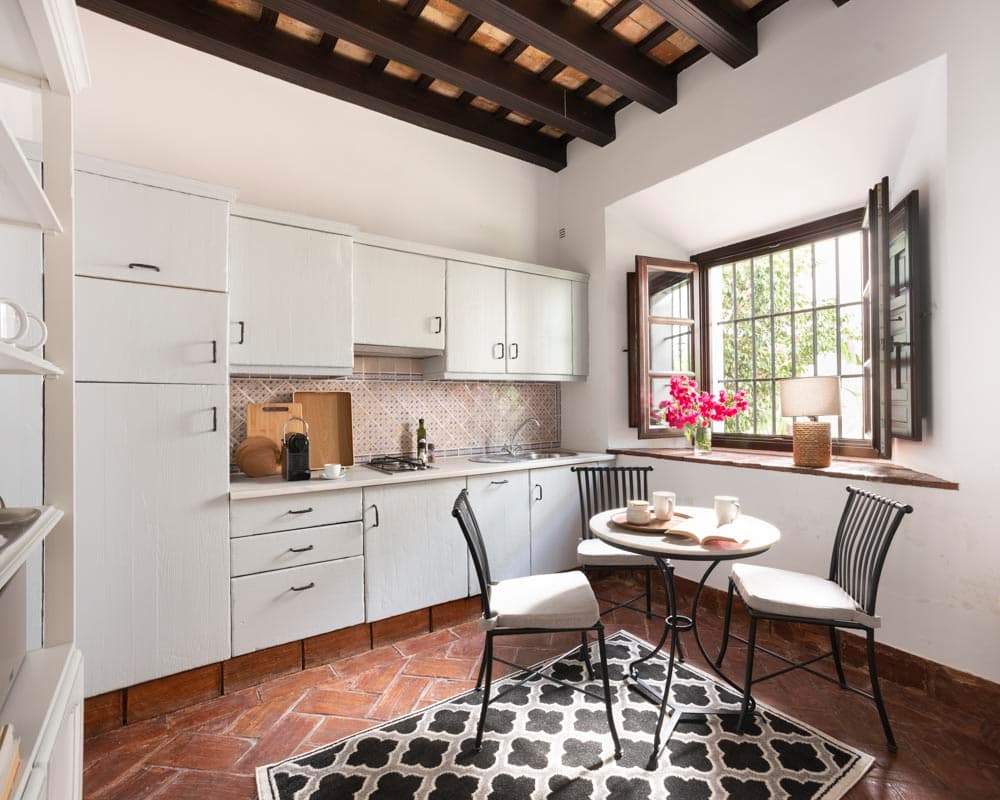 Cocina rústica con muebles blancos envejecidos, alicatado artesanal en azul y blanco, ventana de madera oscura con alféizar de piedra. Mesa redonda de bistró negra con sillas de hierro y cojines blancos, alfombra de geométrica en negro y blanco. Suelo de barro rojo en espiga.