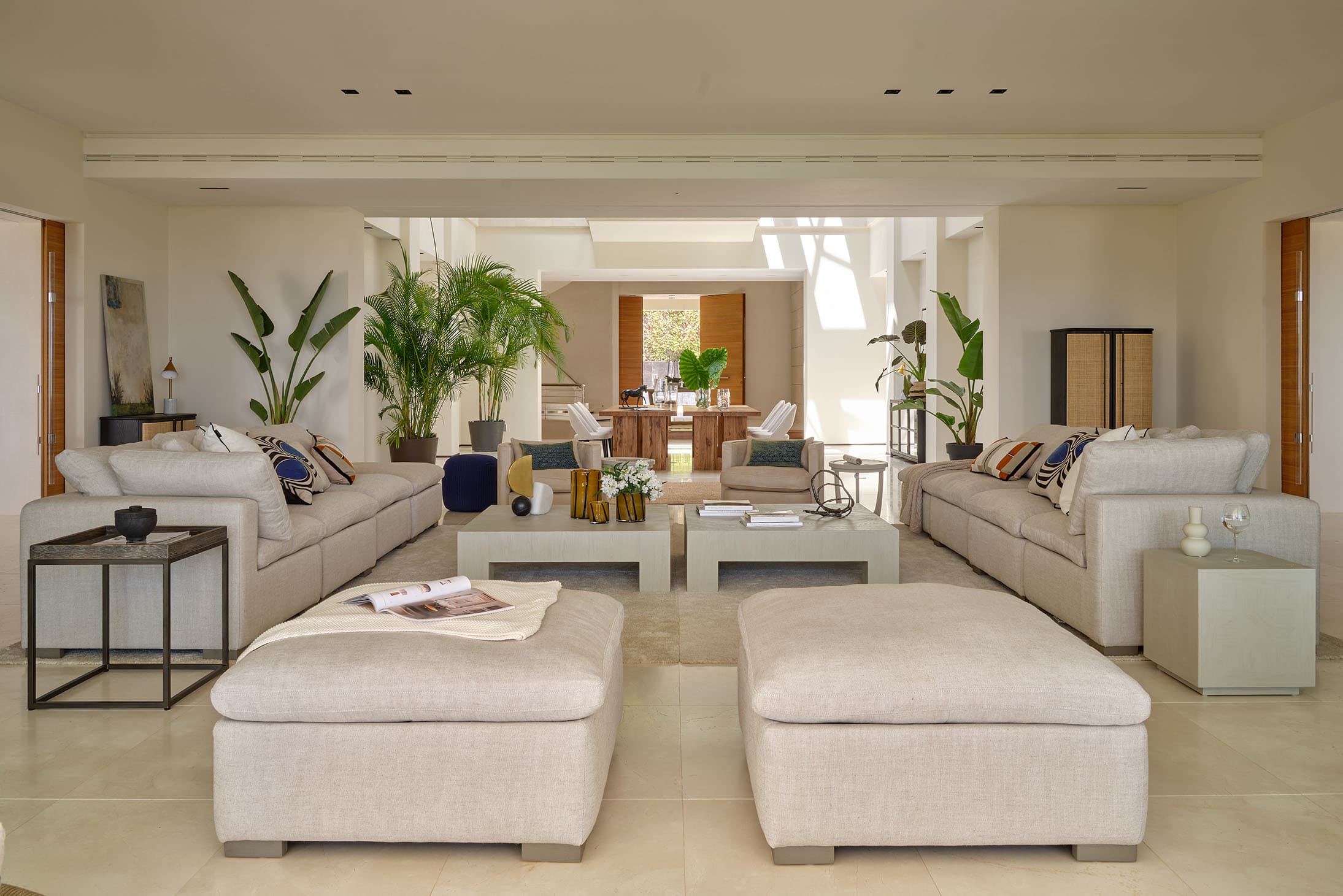 Gran salón de planta abierta con sofás modulares simétricos en beige claro, otomanas dobles, mesas de café bajas y decoración vegetal, que desemboca en el comedor y la cocina.