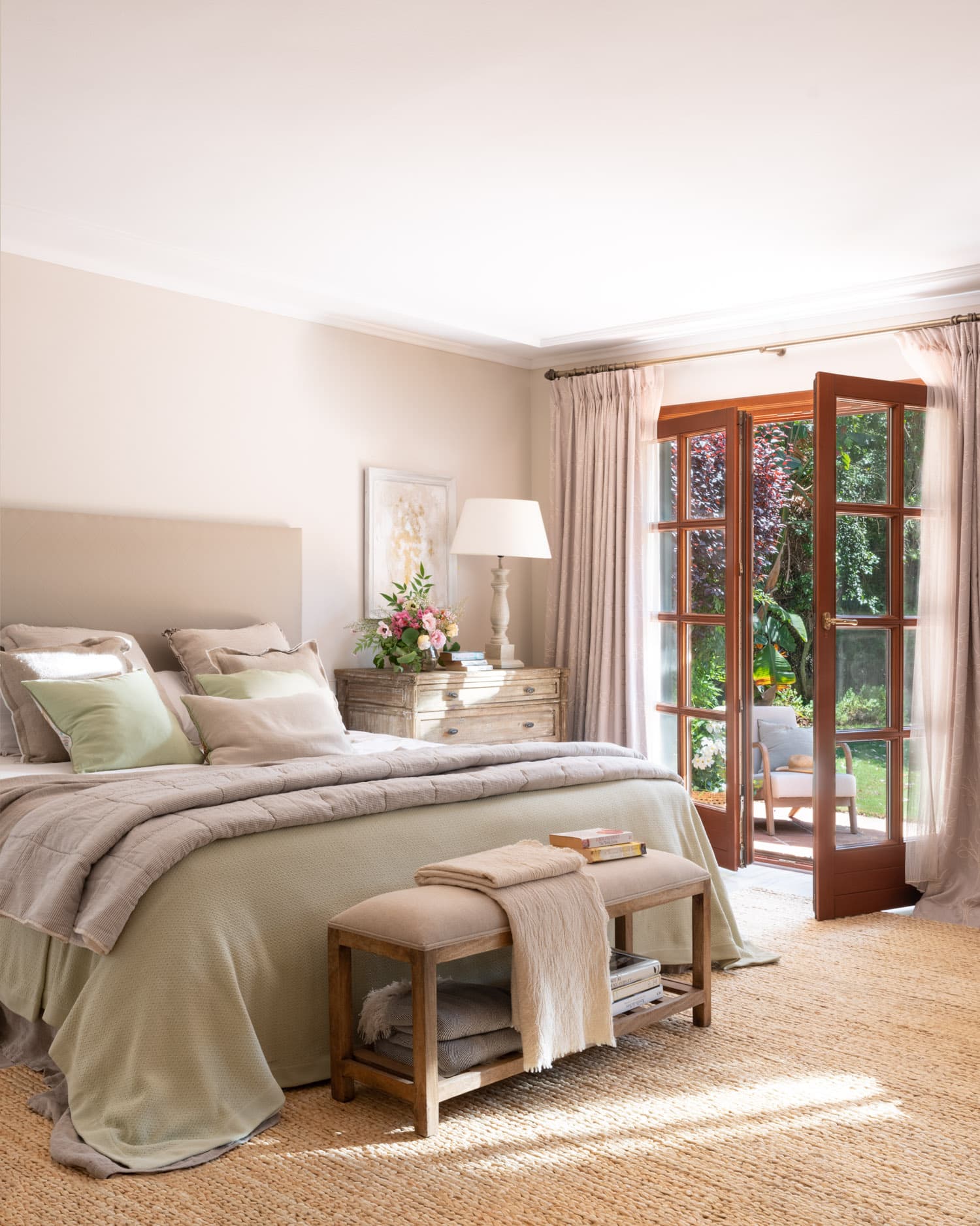 Dormitorio principal de gran calma y luminosidad, con cabecero tapizado en beige y ropa de cama en capas de verde sage, gris y lino. Una mesita de noche provenzal de madera envejecida con lámpara blanca y flores rosas flanquea la cama. La puerta francesa de madera abierta y las cortinas de lino melocotón integran el jardín en la atmósfera del dormitorio. Alfombra de sisal sobre suelo de parqué.