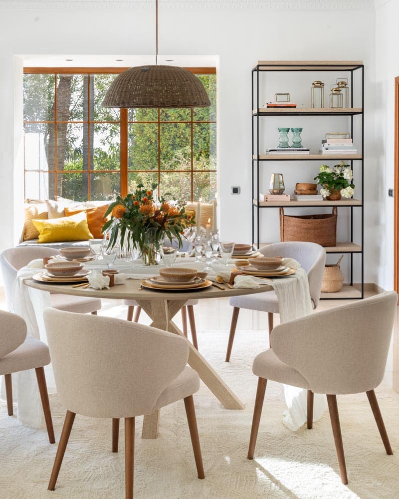 Comedor con mesa redonda y sillas en tono crudo, decorada con una estantería de estructura metálica y baldas de madera llena de libros y objetos decorativos.
