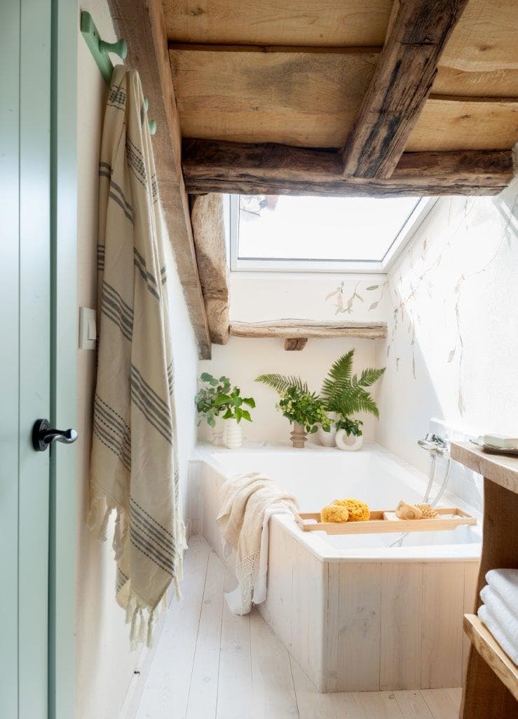 Bañera empotrada en la buhardilla iluminada por una claraboya, con vigas de madera rústica, plantas de helecho y mural de hojas en la pared blanca inclinada. La puerta en verde menta, la toalla a rayas y la bandeja de madera crean un ambiente spa natural.