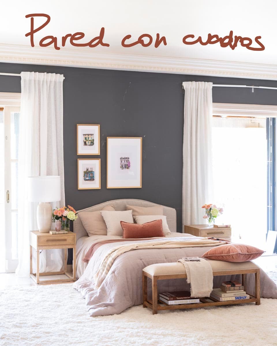 Fotografía de referencia con anotación 'Pared con cuadros' sobre el dormitorio principal, mostrando la cama frente a la pared gris con una composición de tres cuadros enmarcados en dorado con ilustraciones arquitectónicas, mesitas con lámparas blancas y baúl tapizado al pie.