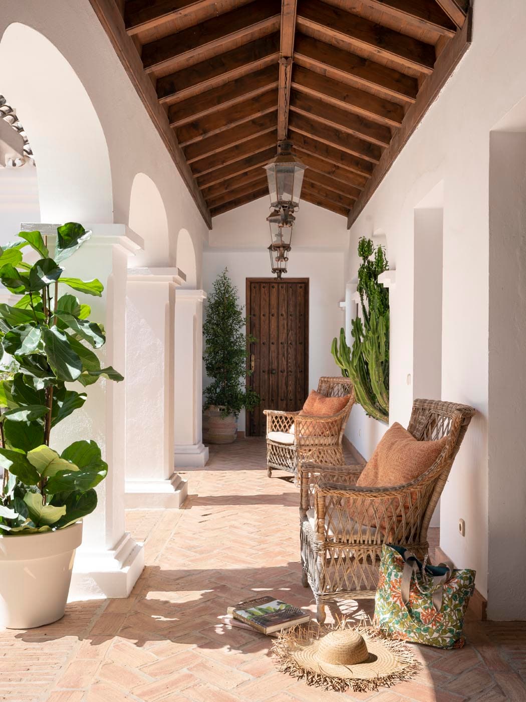 Porche de entrada con bóveda de madera y arcos blancos encalados, donde sillas de mimbre y plantas tropicales crean una bienvenida cálida y mediterránea. El suelo de barro en espiga y la lámpara colgante de latón refuerzan el carácter rústico de lujo de la villa.
