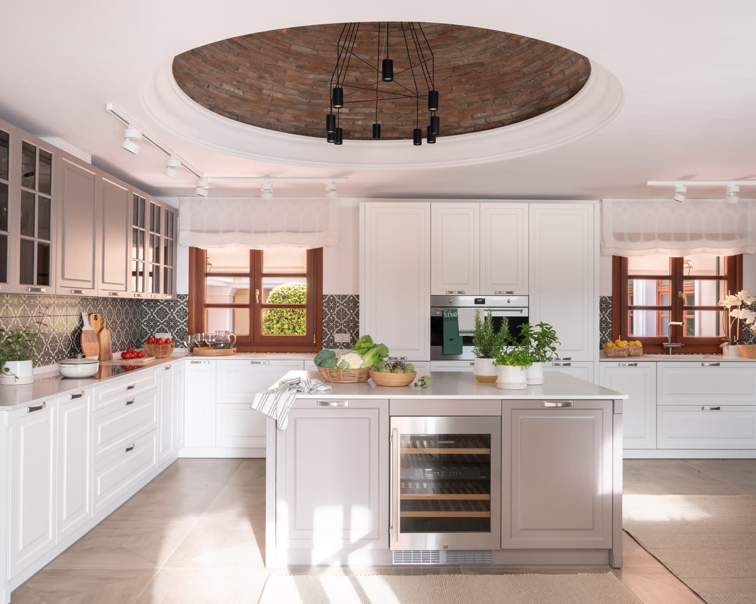 Cocina de gran amplitud con muebles lacados en blanco, encimeras de cuarzo claro y un llamativo backsplash de azulejo hidráulico en negro y blanco. Una cúpula circular en el techo, revestida de ladrillo visto antiguo, acoge una corona de focos negros. La isla central con bodega integrada y vegetales frescos sobre la encimera remite a una cocina de trabajo activa y elegante.