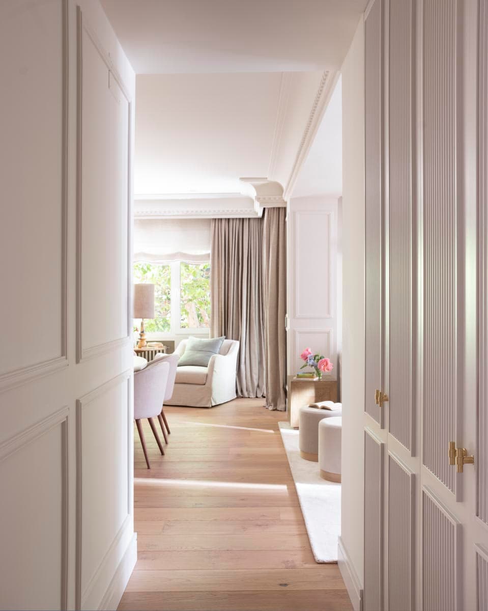 Pasillo central del piso con armarios empotrados de frente estriado en blanco a ambos lados con tiradores de latón dorado, suelo de roble natural y molduras clásicas en techo. Al fondo se ve la estancia de lectura con sillón tapizado, cortinones de lino y la luz natural que entra por los ventanales.