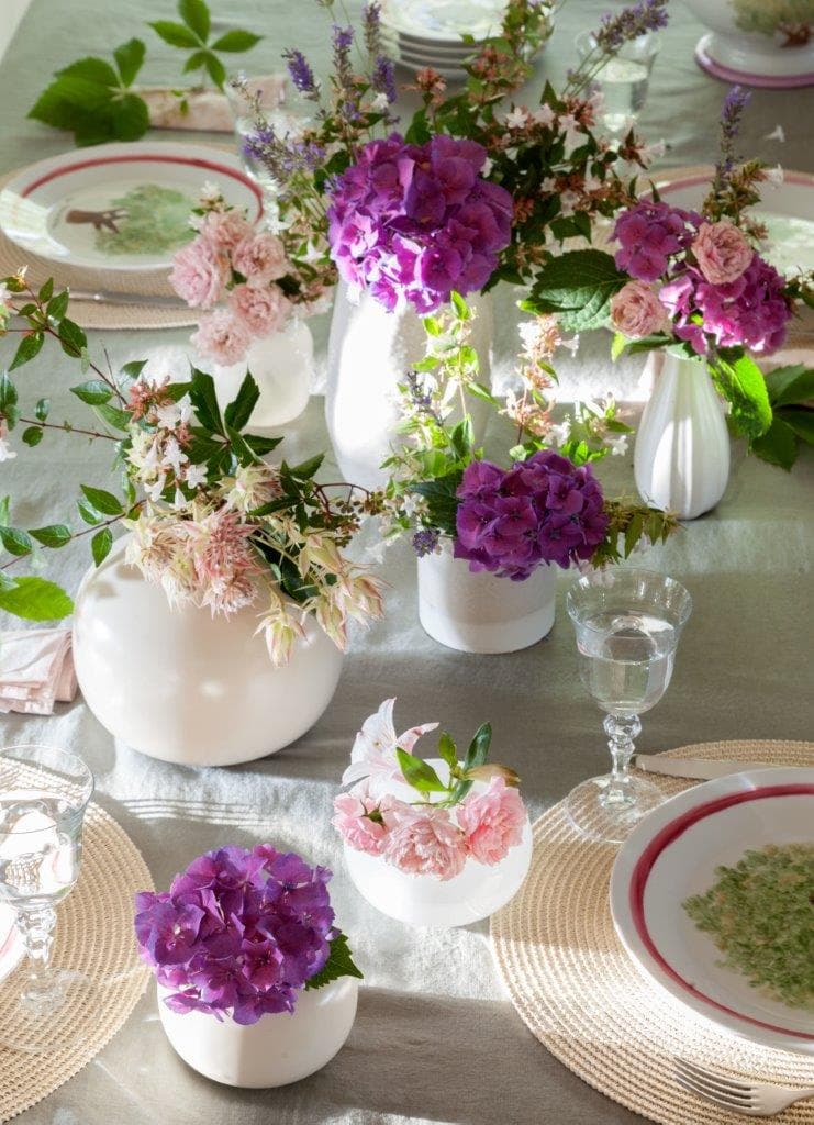 Detalle cenital de una mesa de comedor con camino de lino gris y múltiples jarrones blancos con flores silvestres: hortensias moradas, rosas de jardín, clemátide y lavanda. Salvamanteles de esparto y platos con motivos de árbol en verde, borde rojo y copas de cristal soplado. Una composición floral campestre de gran delicadeza.