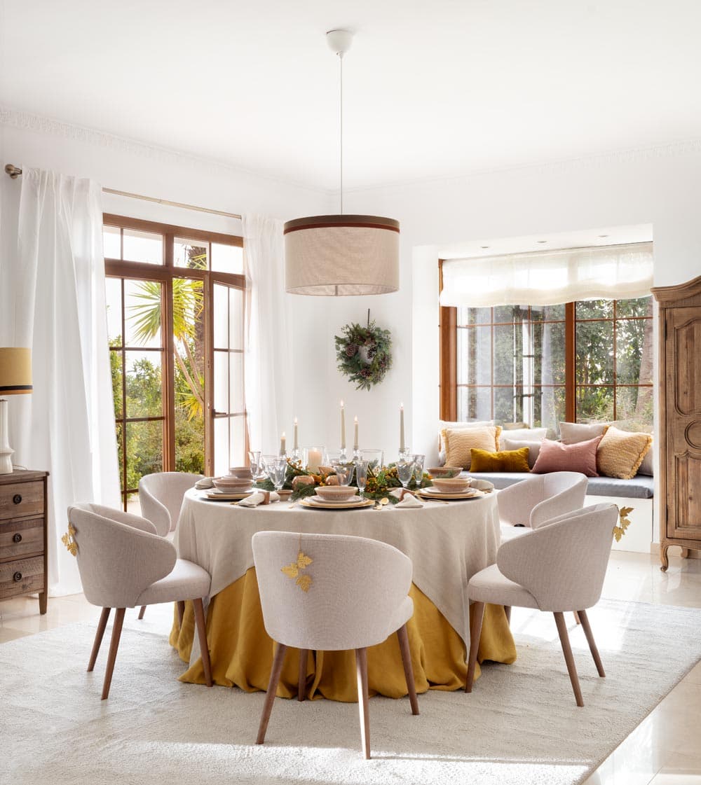 Comedor luminoso con mesa redonda vestida con mantel en lino crudo sobre falda en mostaza viejo, rodeada de sillas tapizadas en bouclé beige con detalles en dorado. La decoración navideña incluye corona de pino natural, velas y vajilla artesanal sobre un fondo de ventanas con carpintería en madera.