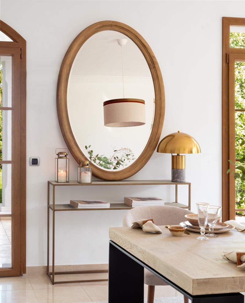 Detalle decorativo con gran espejo oval de madera natural sobre una consola de estructura metálica, acompañado de una lámpara de mesa en cobre dorado y velas en portavelas de cristal.