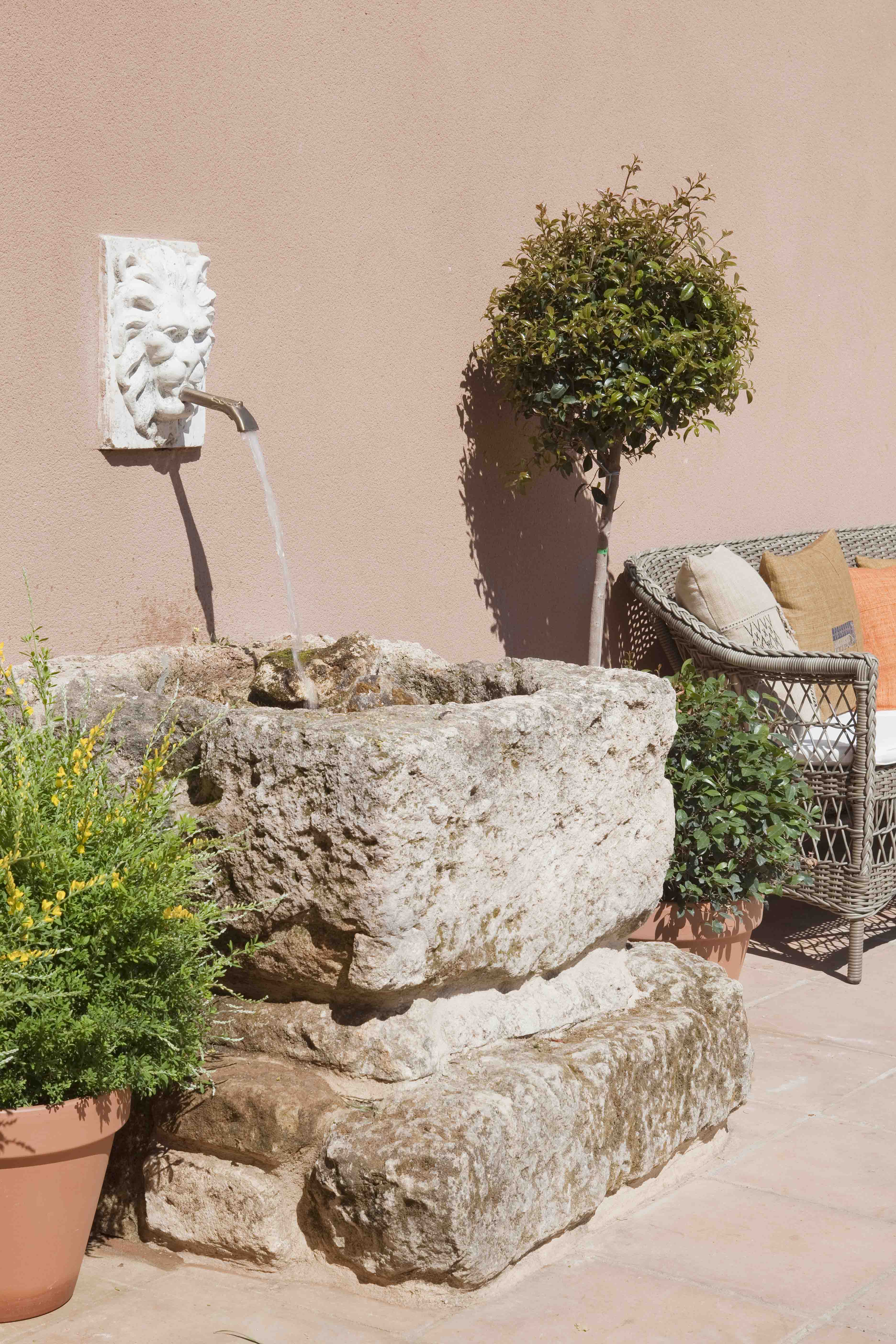 Rincón de terraza con una fuente de piedra muy antigua con mascarón de león de mármol blanco. Junto a ella, macetas de barro con plantas aromáticas y una silla de ratán con cojines. Un árbol podado en forma de bola aporta verticalidad al conjunto.