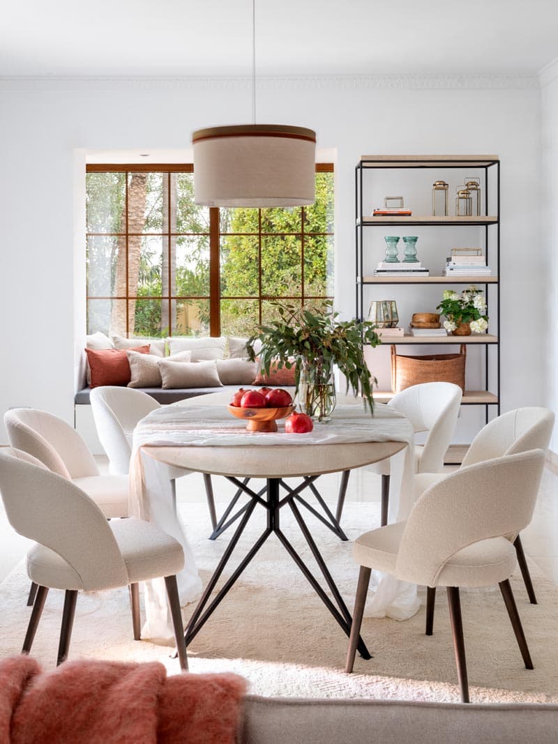 Mesa de comedor redonda con base de patas negras, centrada con granada y flores silvestres otoñales, sillas tapizadas en blanco y una lámpara de tambor colgante bajo luz natural. La estantería industrial al fondo y el banco-ventana con cojines coral completan este comedor de inspiración contemporánea.