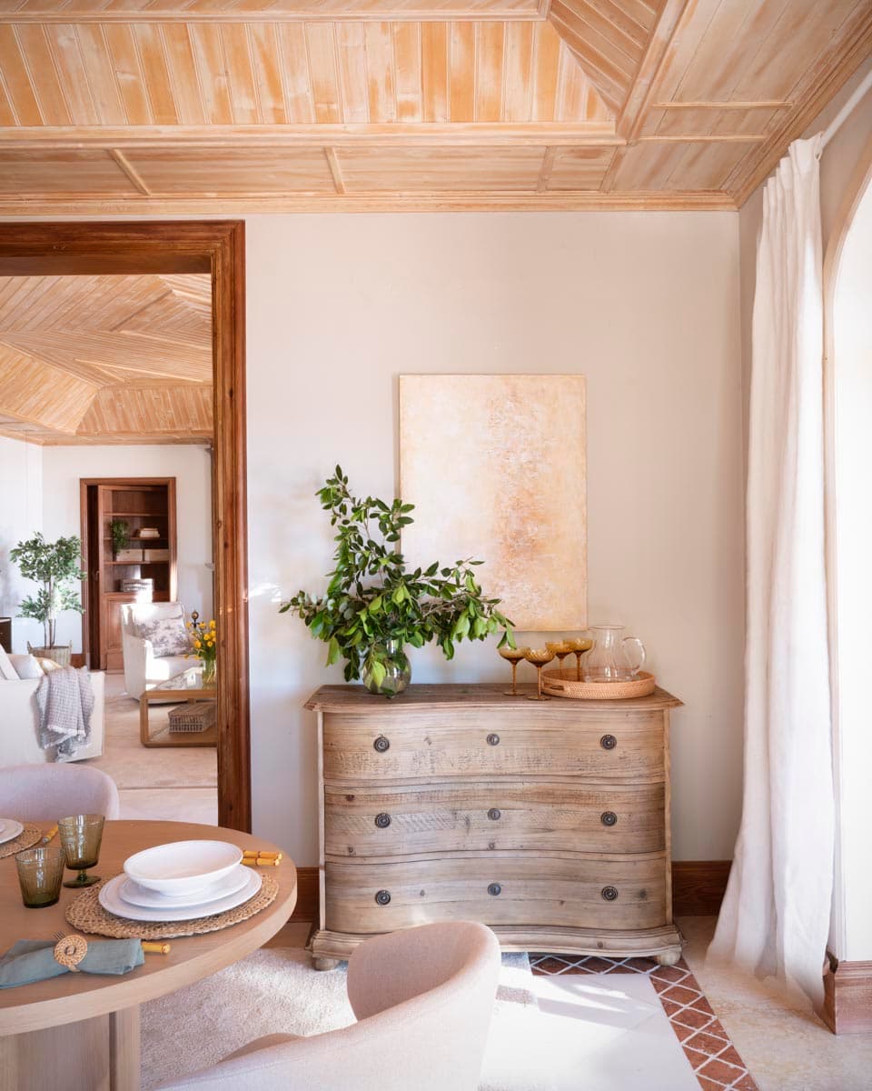 La cómoda gustaviana de madera decapada junto al acceso al comedor, decorada con una bandeja de ratán con copa de cristal ámbar y jarra, un centro de ramas con brotes blancos y un cuadro abstracto en tonos dorados y beige. El techo artesanal y el suelo de mármol con cenefa enmarcan la escena.