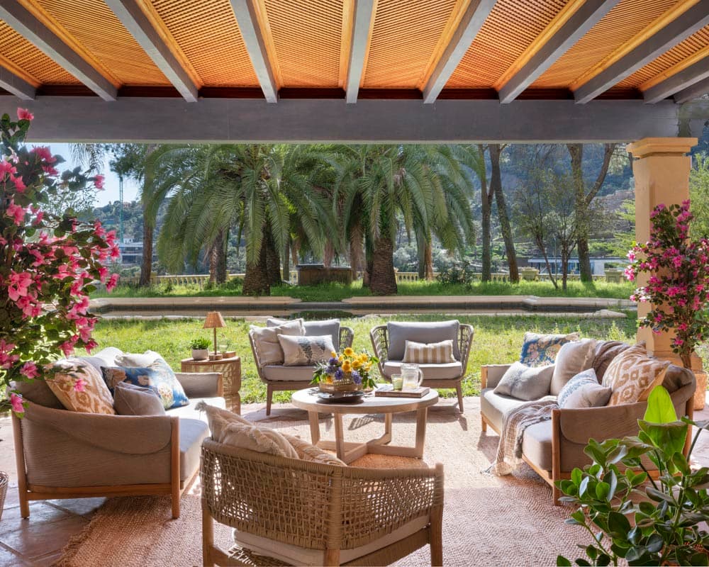Amplio porche cubierto con techo de caña sobre vigas de madera, zona de estar con sofás y sillones de ratán con cojines en gris y tonos azul índigo, flanqueado por buganvillas en flor rosa intenso y con vistas a la piscina y palmeras del jardín.