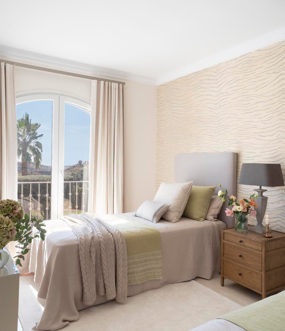 Dormitorio de invitados luminoso con papel pintado en tonos crema con ondas. Cama individual con cabecero tapizado en gris, ropa de cama en beige y verde pistacho y manta de punto. Mesita de madera con lámpara gris y flores. Puerta de arco con balcón y vistas a palmeras y montañas de la Costa del Sol.