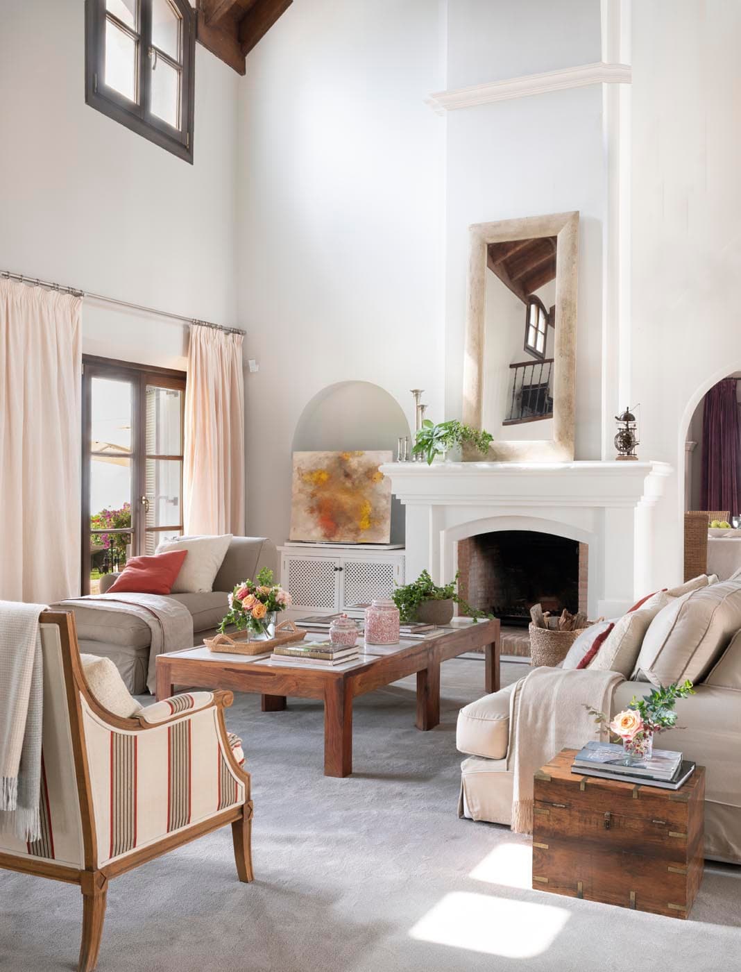 Vista lateral del salón principal que muestra el sillón de rayas en primer plano, la chimenea con espejo de piedra encima y sofás en lino claro alrededor de la mesa de centro de madera. La alta ventana con cortinas en rosa pálido y los detalles florales añaden frescura al ambiente.