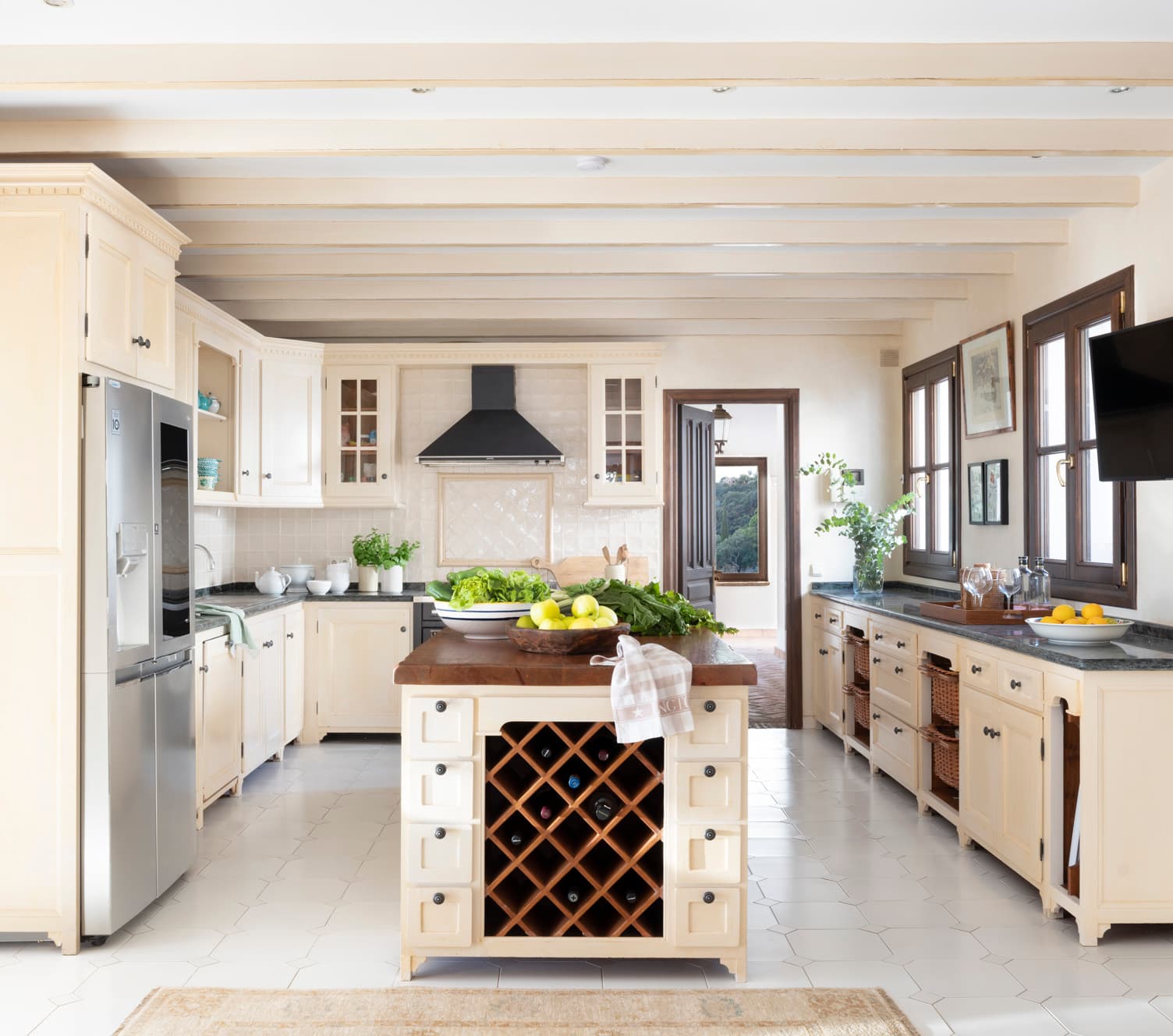 Amplia cocina de estilo clásico con muebles lacados en blanco hueso, vigas de madera en el techo y suelo de baldosa blanca hexagonal. La isla central incorpora una bodega de madera con forma de rombo y superficie de madera oscura. Encimeras de granito gris jaspeado. Ventanas con carpintería oscura enmarcan vistas al jardín.