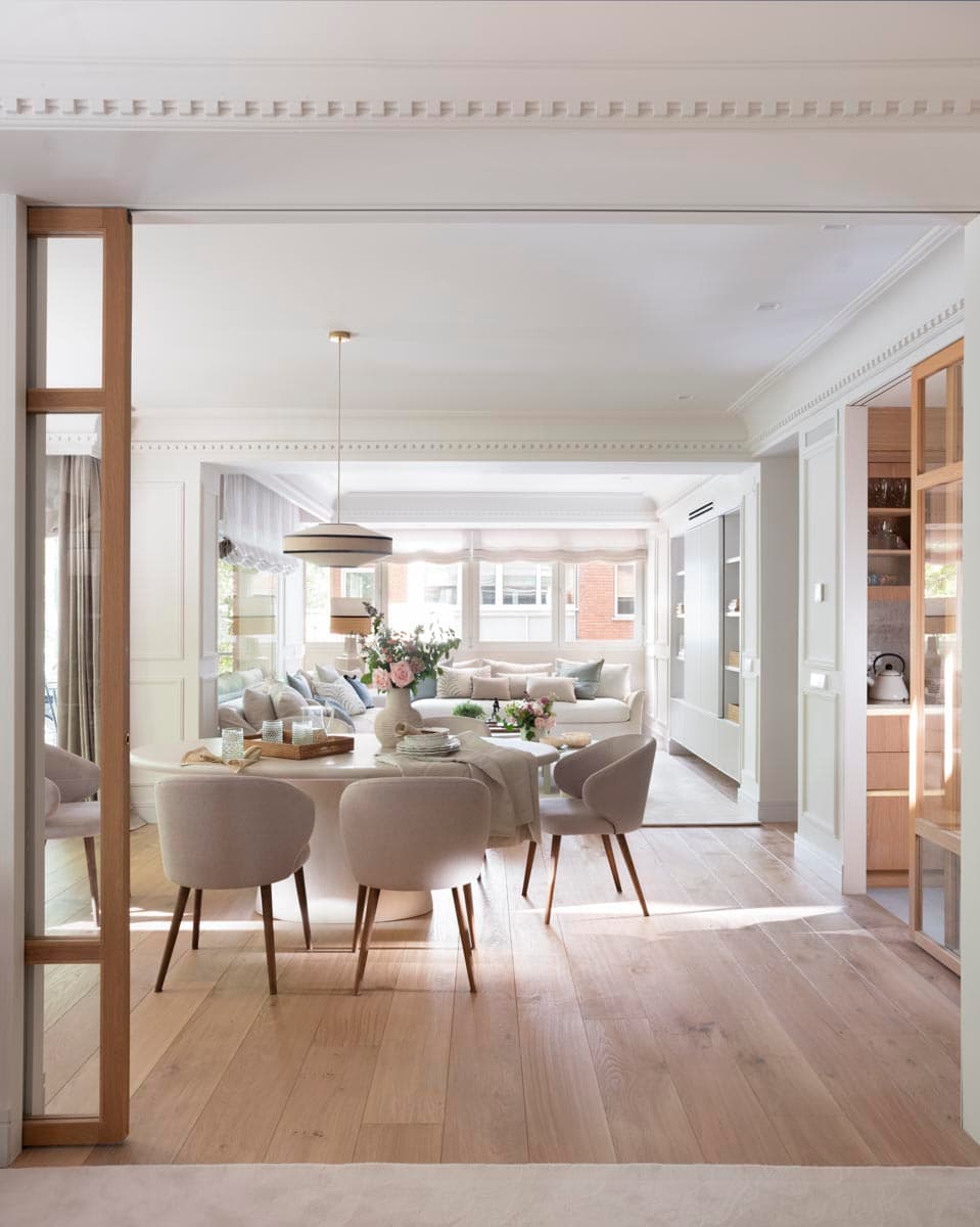 Vista desde el umbral de la entrada enmarcado en madera de roble hacia el comedor, con mesa ovalada blanca y sillas tapizadas en gris perla, y al fondo el salón iluminado por grandes ventanales. A la derecha se atisba la cocina integrada con vidriera de madera y estantería con copas.