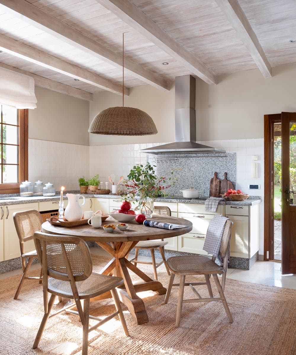 Vista de la cocina desde el ángulo del comedor informal, con la campana extractora metálica como elemento central y fondo de encimera de granito con tabla de cortar, pan artesanal y granadas rojas decorativas. Las sillas de mimbre, el banco de la mesa de madera y la puerta al jardín aportan un aire campestre y luminoso.