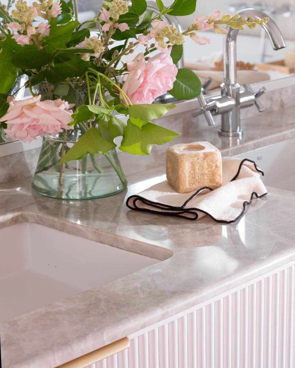 Primer plano del lavabo encastrado en encimera de mármol gris veteado con grifo de pomo cruzado en cromo plateado, jabón artesanal sobre paño de lino con ribete negro y jarrón de cristal con rosas rosas y follaje verde. El mueble bajo con frente estriado en blanco roto y pomo dorado enmarca la composición.