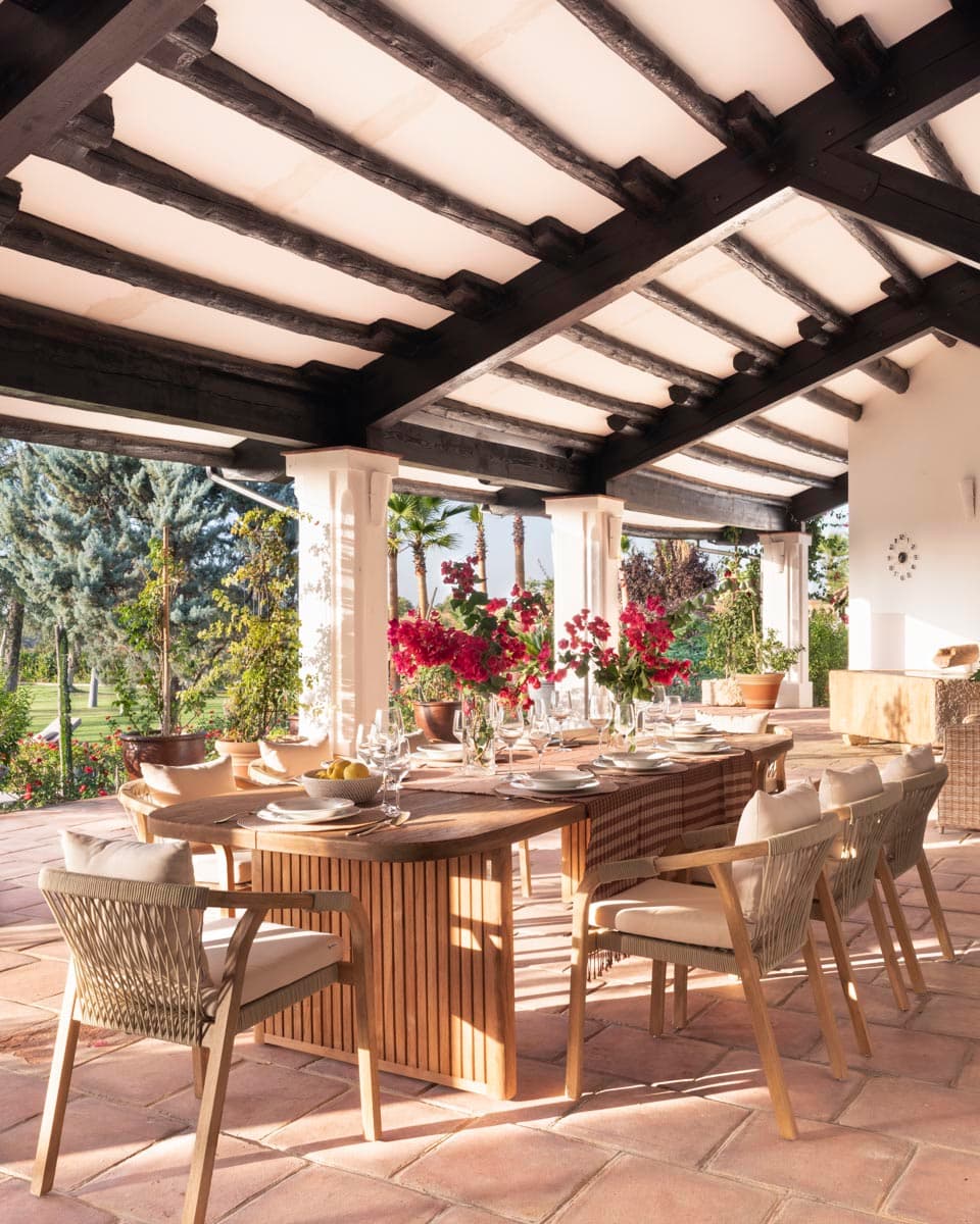 Mesa de comedor exterior ovalada de madera bajo el porche de vigas, con sillas de cuerda, vajilla puesta para la celebración y bougainvilleas rojas en plena floración al fondo.