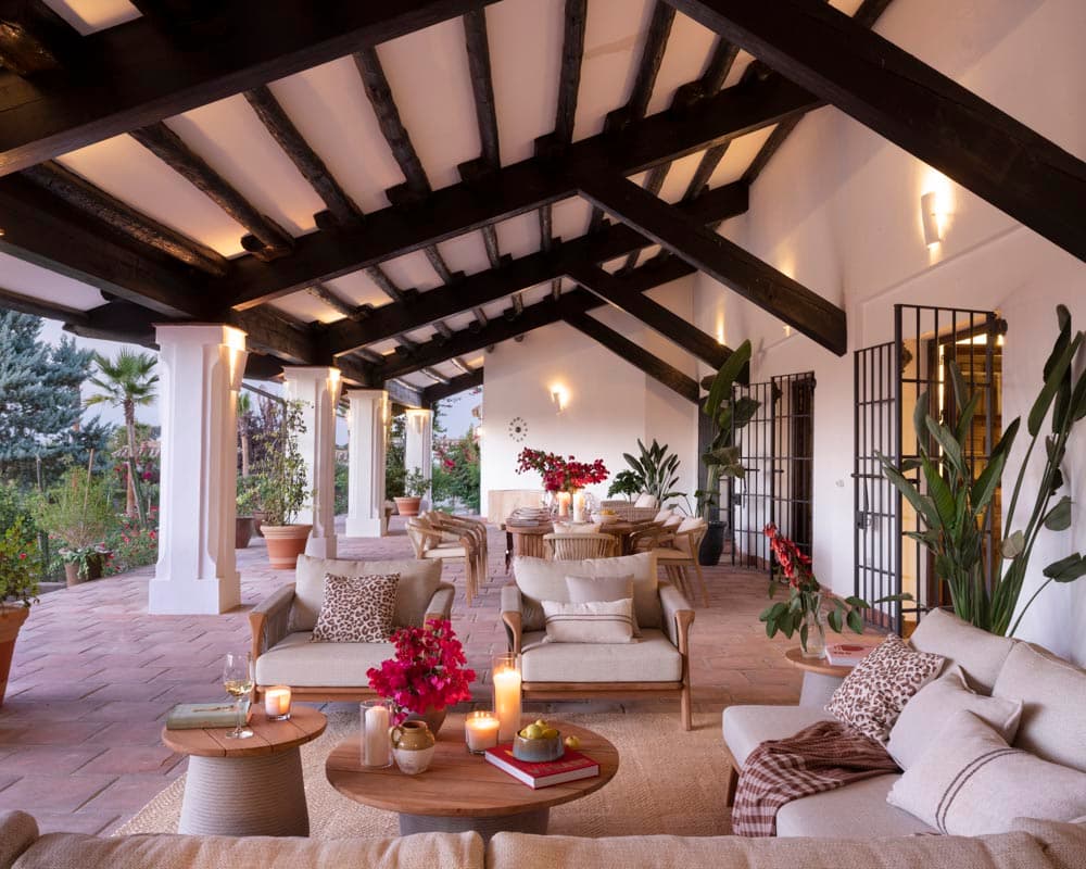 Terraza cubierta iluminada al anochecer con vigas de madera negra y columnas blancas. Zona de sofás con cojines blancos y estampado leopardo, apliques de pared y velas. Al fondo, zona de comedor exterior con buganvilla y plantas tropicales.