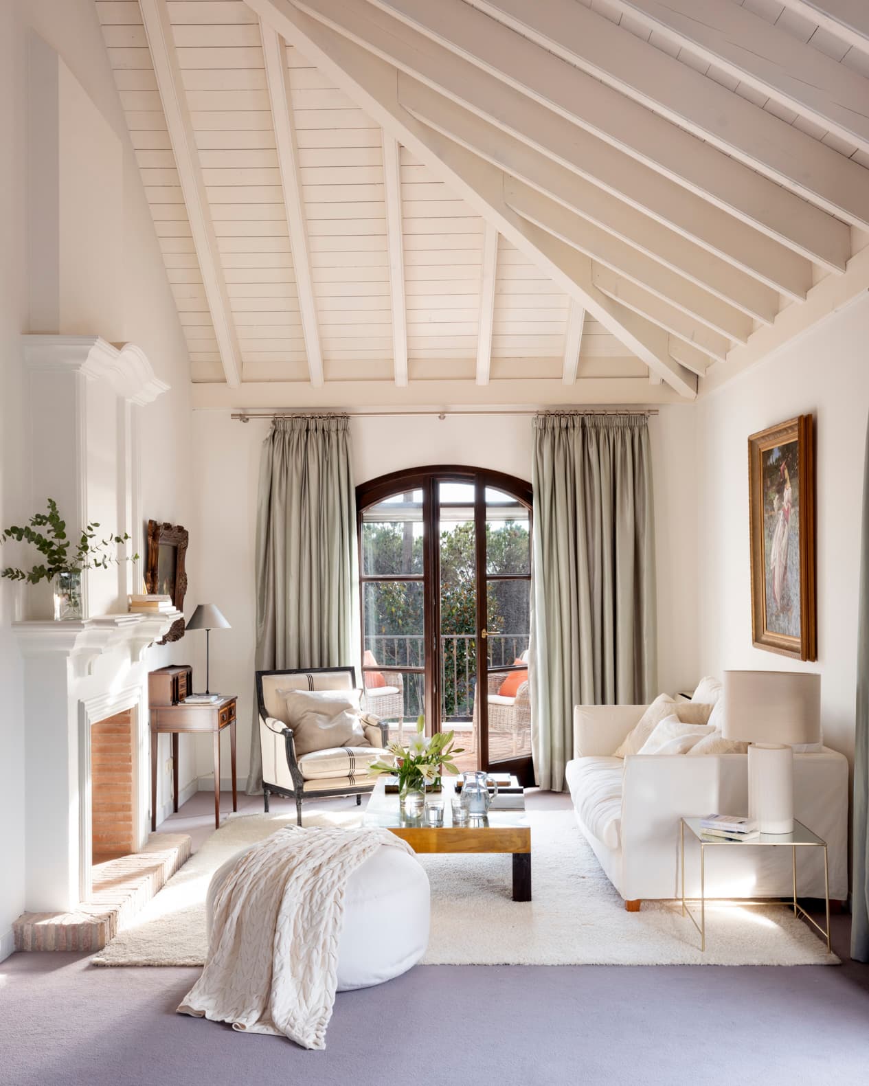 Sala de estar con espectacular cubierta a dos aguas blanca y vigas vistas. La paleta es totalmente blanca con sofá de lino, puf redondo, chimenea de mármol blanco y cuadro clásico. Cortinas verde grisáceo flanquean la puerta arquitrabada que da al balcón. La iluminación natural y la sencillez crean un ambiente sereno.
