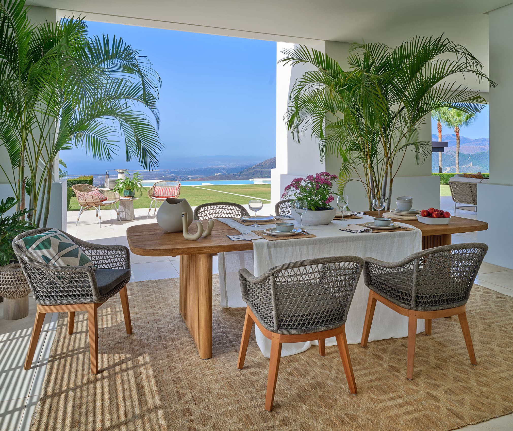 Comedor de terraza con mesa de teka y sillas trenzadas, rodeado de palmeras, con vistas a la piscina de borde infinito y al mar Mediterráneo en el horizonte.