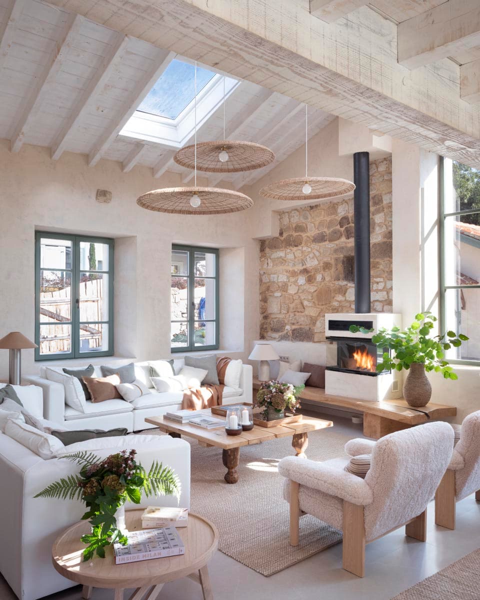 Salón de doble altura con lucernario, vigas de madera blanqueadas y chimenea integrada en muro de piedra vista, decorado con sofás blancos, lámparas de ratán planas y mesa de centro de madera rústica.