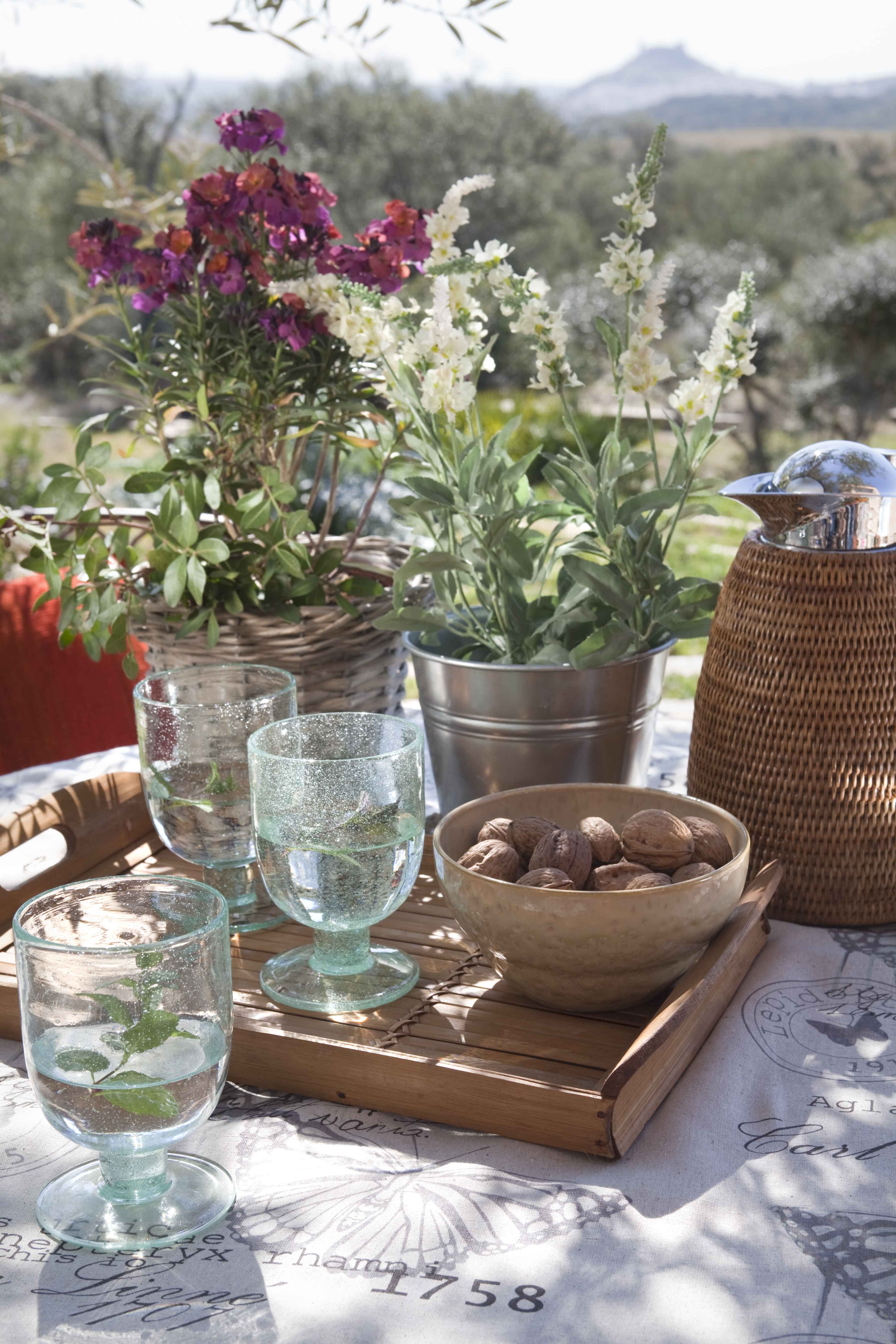 Detalle de una mesa en la terraza exterior con copas de agua artesanales, flores silvestres en macetas metálicas, un cuenco de madera con frutos secos y una cesta de mimbre. Al fondo, un paisaje de campo abierto con sierra en el horizonte bajo un cielo despejado.